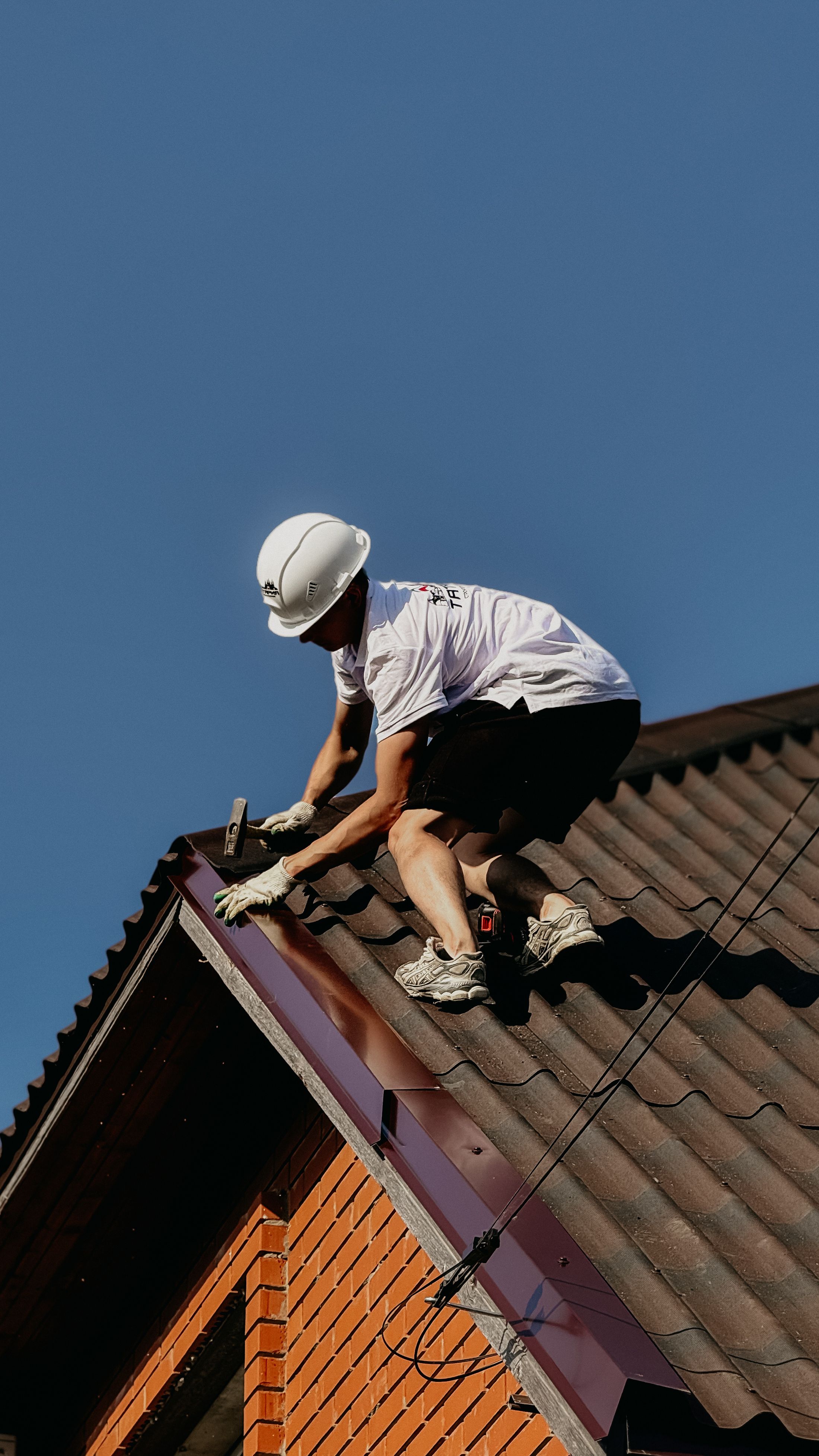 Dachdecker mit Helm verlegt Ziegel am First eines Steildachs vor blauem Himmel